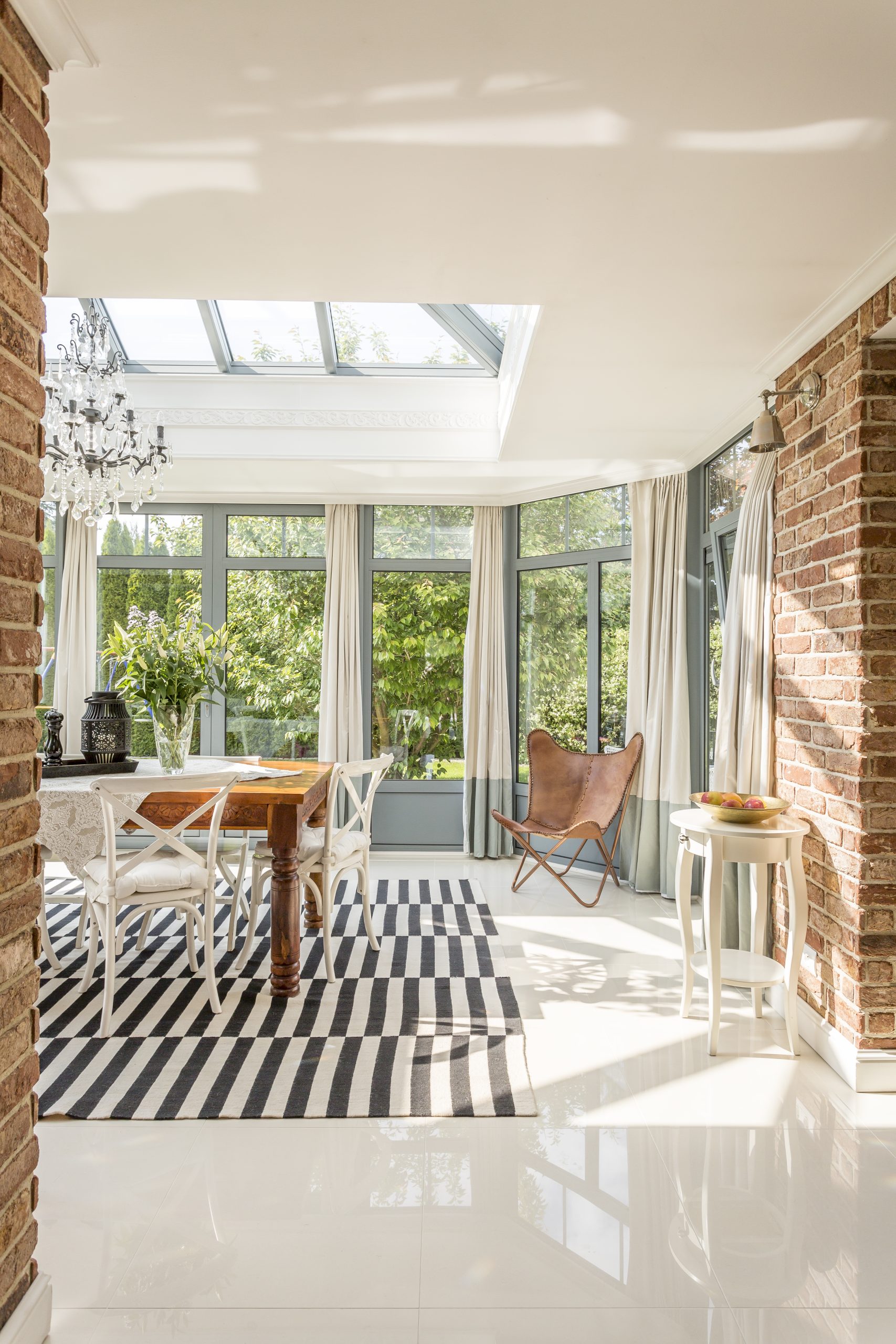 Spacious, modern dining room with windows around, glazed roof, stylish chandelier, brick walls, stripy carpet and table with chairs in the middle