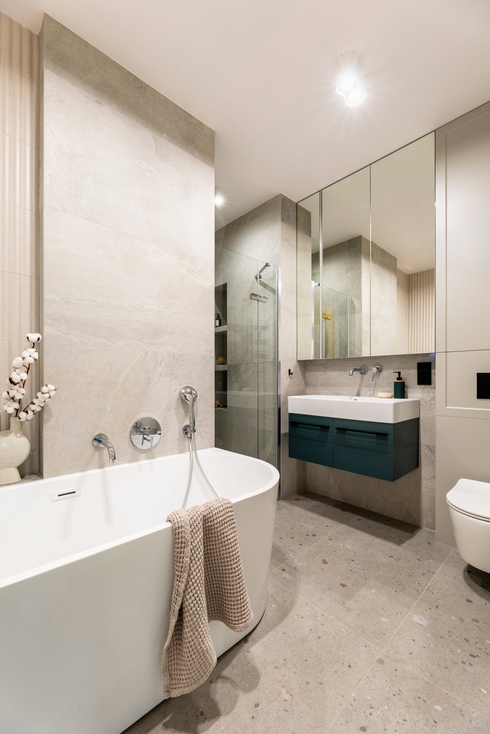 Modern bright bathroom interior with lamella wall. Big white bath with silver faucet and brown towel. Washbasin with dark cabinet and morror.